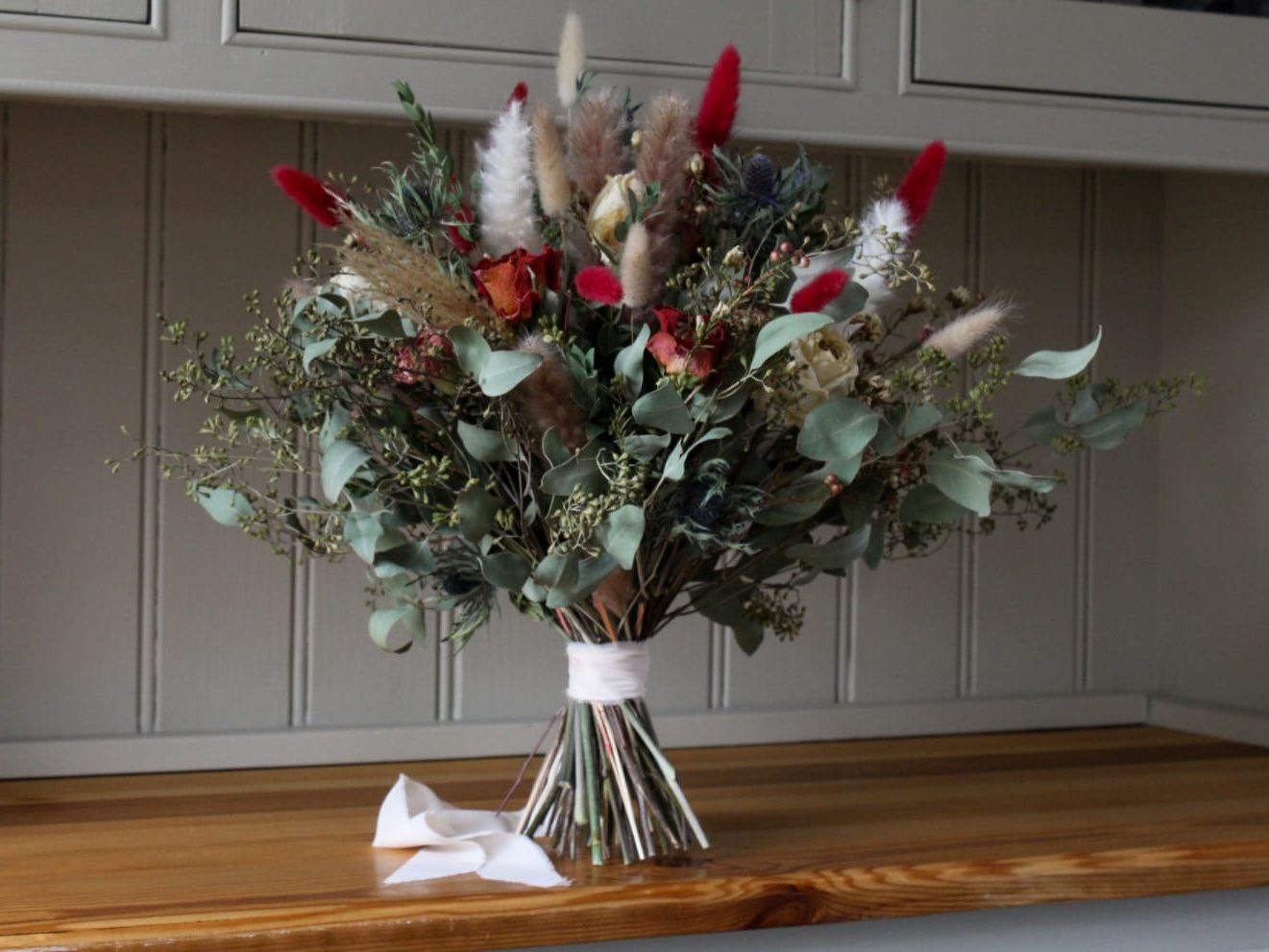 Wedding bouquet featuring dried grasses, orange and cream flowers and eucalyptus foliage tied with ribbon on a wooden surface