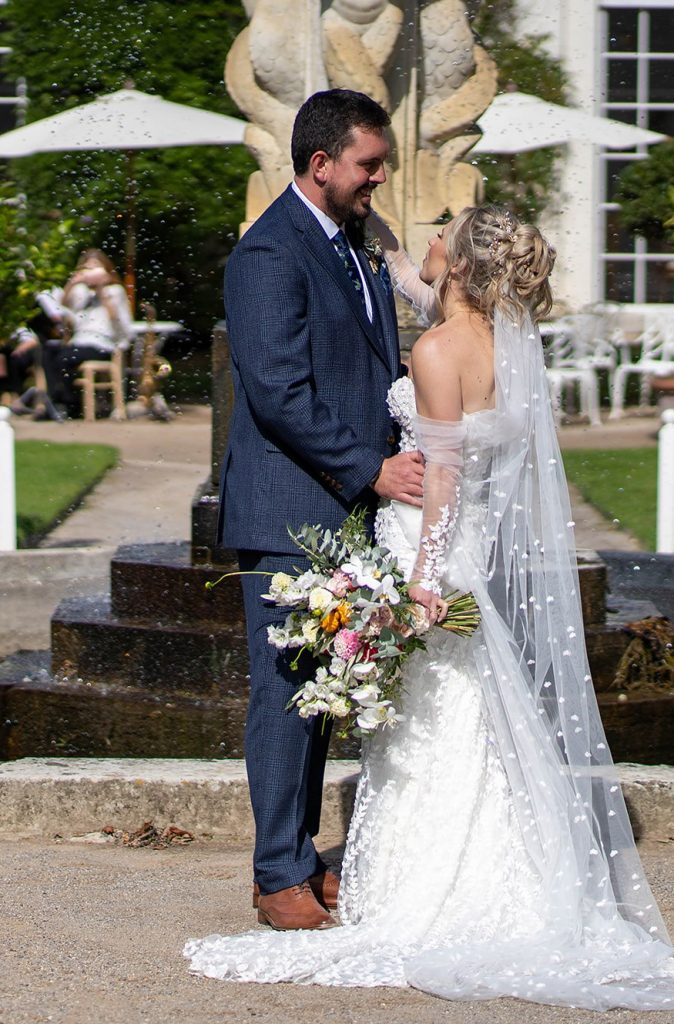 Bride and groom smiling stood near water fountain with bespoke wedding bouquet by The Flowers of May, Cornwall wedding florist