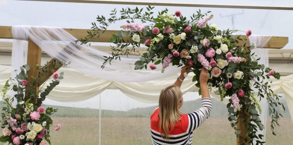 Florist arranging a floral arch with pink and white seasonal blooms and greenery by The Flowers of May, Cornwall wedding florist