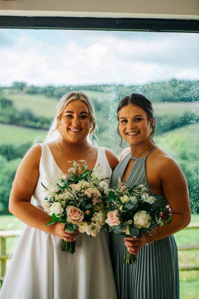 Bride and bridesmaid holding bouquets with mixed seasonal blooms and greenery by The Flowers of May, Cornwall wedding florist
