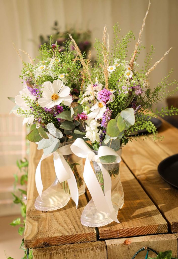 Natural wildflower table arrangements in clear kids welly boots with white ribbons, featuring daisies, purple blooms and greenery