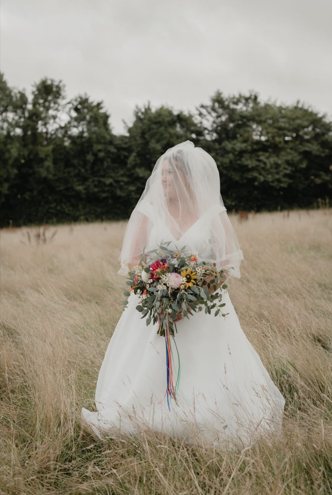 Bride in white dress standing in a grassy field holding a seasonal wedding bouquet with mixed foliage and colourful blooms