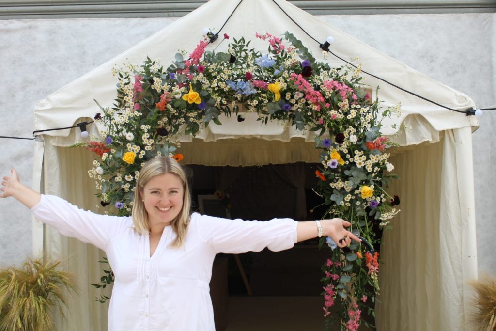 Wedding floral arch display with mixed bright seasonal blooms and greenery by The Flowers of May, Cornwall wedding florist. Megan Haigh, florist of The Flowers of May smiling in front of the archway.